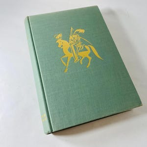 May include: A hardcover book with a light green cover. A gold illustration of a knight on horseback is centered on the front. The book's spine is visible, and the pages are closed. The book is on a white surface.