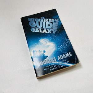 Könnte beinhalten: Ein Taschenbuch von "Per Anhalter durch die Galaxis" von Douglas Adams. Der Umschlag zeigt einen blauen und schwarzen Sternenhimmel mit einem Raumschiff und einer Gruppe von Menschen, die auf einem Planeten stehen. Das Buch ist eine Film-Ausgabe mit einem Nachwort von Robbie Stamp.