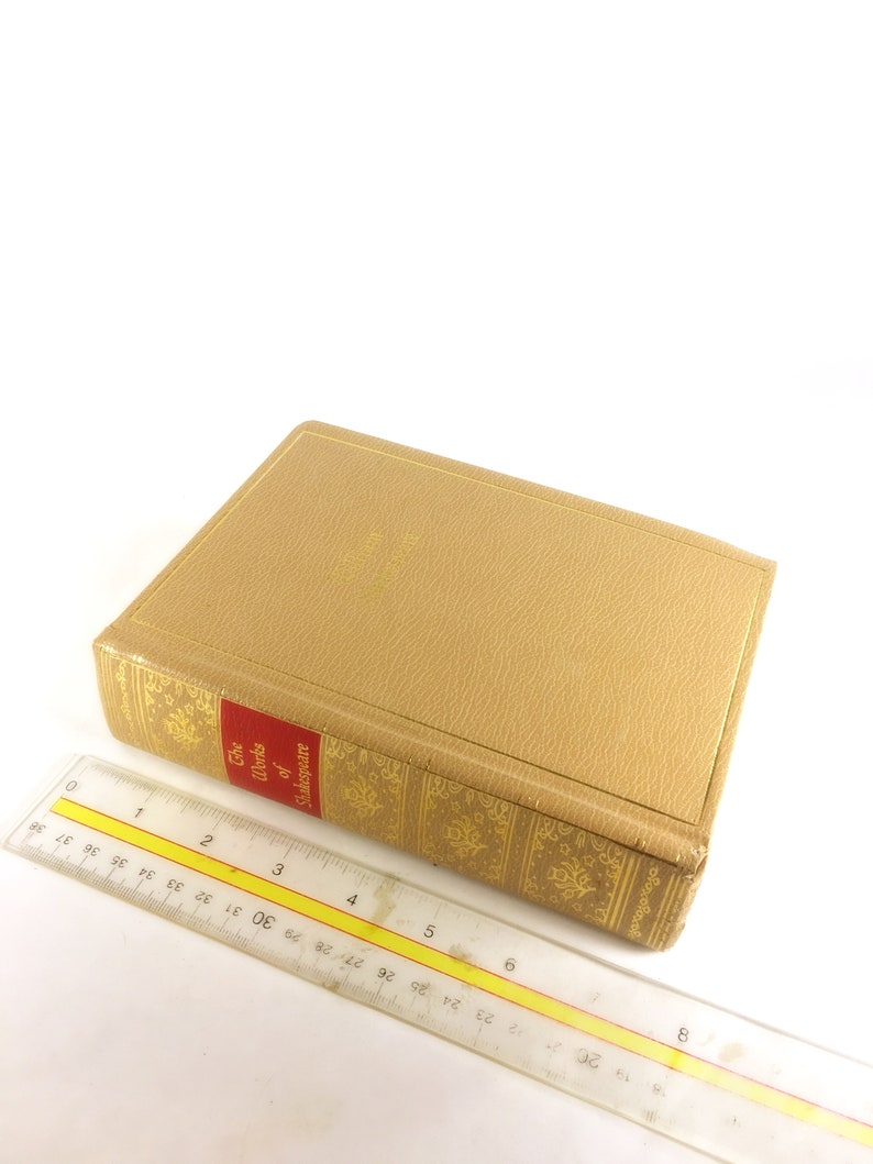 May include: A hardcover book titled "The Works of Shakespeare" with a tan cover and gold embossed details. A clear ruler is placed next to the book, showing measurements in inches.