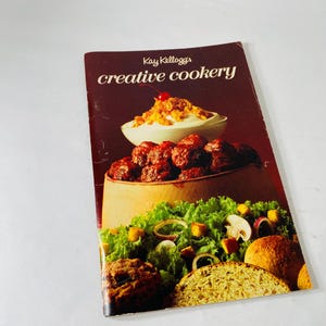 Könnte beinhalten: Vintage Kochbuch mit dem Titel "Kay Kellogg's creative cookery" mit dunkelrotem Einband. Das Cover zeigt eine Lebensmittelillustration von Fleischbällchen, Salat und Brot. Das Kochbuch ist leicht abgenutzt.