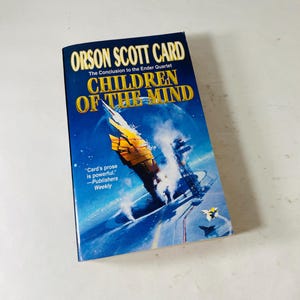 Könnte beinhalten: Ein Taschenbuch mit dem Titel "Children of the Mind" von Orson Scott Card. Das Cover zeigt ein futuristisches Raumschiffdesign in Gelb und Orange vor blauem Hintergrund. Das Buch ist Teil der Reihe "The Ender Quartet".