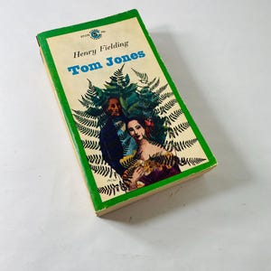 Puede incluir: Un libro de bolsillo vintage titulado "Tom Jones" de Henry Fielding. La portada presenta un borde verde, un fondo crema y una ilustración de un hombre y una mujer rodeados de helechos. Una novela clásica.