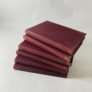 May include: A stack of six burgundy-colored books with gold lettering. The titles are visible on the spines, including "The Winter's Tale" and "Much Ado About Nothing." The books are stacked at a slight angle.