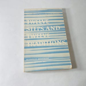 Puede incluir: Un libro antiguo titulado "Twelve Steps and Twelve Traditions" de Alcohólicos Anónimos. La portada presenta rayas horizontales azules y texto blanco. El título del libro se muestra de forma destacada, con el nombre del autor en la parte inferior.