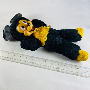 May include: A black and yellow plush Mickey Mouse toy with a big smile and large eyes. The toy is lying on its back with its arms outstretched.