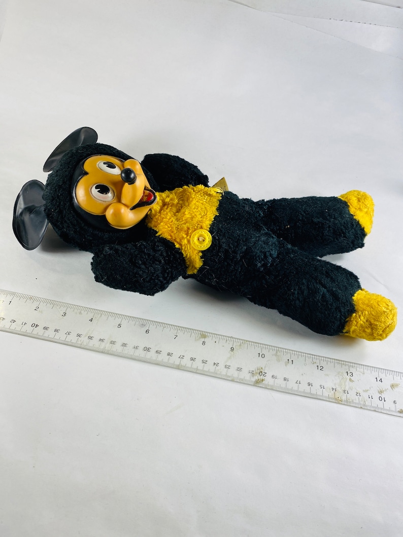 May include: A black and yellow plush Mickey Mouse toy with a big smile and large eyes. The toy is lying on its back with its arms outstretched.