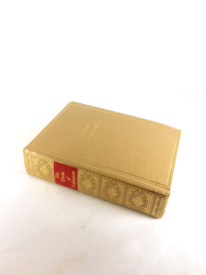 May include: A hardcover book titled "The Works of Shakespeare." The book has a tan cover with gold lettering and decorative patterns. The spine features a red label with white text. The book is closed and resting on a white surface.