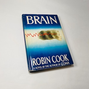 May include: A blue paperback book titled "Brain" by Robin Cook, a novel by the author of "Coma". The cover features a woman's head with a red wavy line above it.