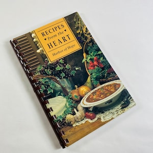 May include: A spiral bound cookbook titled "Recipes from the HEART Harbor of Hope" with a colorful image of a bowl of stew, vegetables, and fruit on the cover.