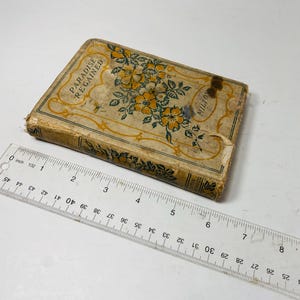 May include: An antique book titled "Paradise Regained" by Milton. The book has a worn, aged cover with floral and leaf designs in yellow and green. A clear ruler with inch measurements is placed next to the book.