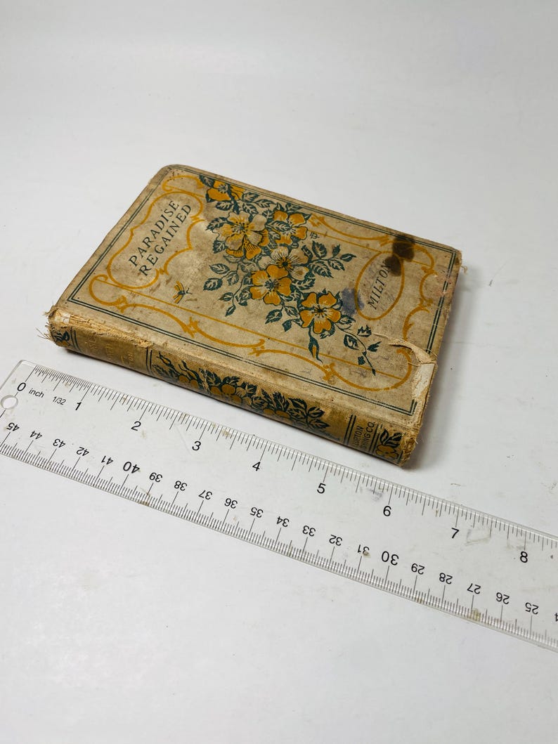 May include: An antique book titled "Paradise Regained" by Milton. The book has a worn, aged cover with floral and leaf designs in yellow and green. A clear ruler with inch measurements is placed next to the book.