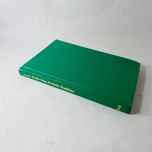 May include: A green hardcover book titled "Al-Anon's Twelve Steps & Twelve Traditions" with gold lettering. The book is closed and lying flat, with the spine visible. The book has a B-8 marking on the bottom right corner.