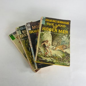 May include: A stack of vintage paperback books by Edgar Rice Burroughs. The covers feature illustrations of jungle scenes and titles like "The Land of Hidden Men" and "The Moon Men." The books are in various shades of green, yellow, and brown.