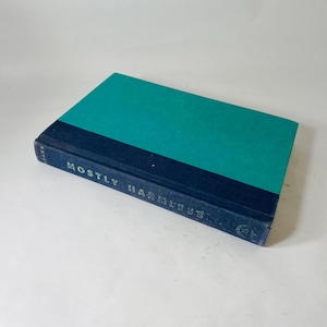 Könnte beinhalten: Ein gebundenes Buch mit türkisgrünem Einband und marineblauem Buchrücken. Der Titel "Mostly Harmless" ist in Silber gedruckt. Das Buch liegt auf einer weißen Oberfläche. Abmessungen nicht sichtbar.