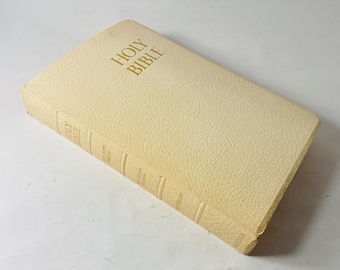May include: A cream-coloured Holy Bible with gold lettering on the cover. The book has a textured cover and is slightly angled, showing the spine with additional text. The edges of the pages are visible.