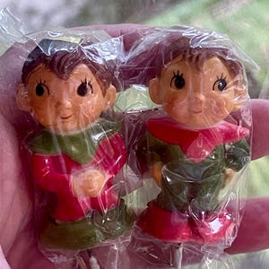 May include: Two small elf figurines encased in clear plastic packaging. Each elf has brown hair, large eyes, and a pointed collar. The left elf wears a red outfit with a green collar and base, while the right elf wears a green outfit with a red collar.