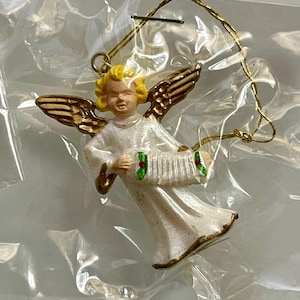 May include: A small, vintage Christmas ornament in the shape of an angel. The angel is wearing a white robe with gold trim and has gold wings. It is holding an accordion and has yellow hair. The ornament is suspended by a gold string.
