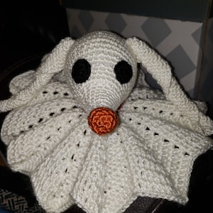 May include: A handmade crochet plush toy of Zero from The Nightmare Before Christmas. The white ghost dog has black button eyes, a small orange nose, and long floppy ears. The plush is resting on a dark surface.
