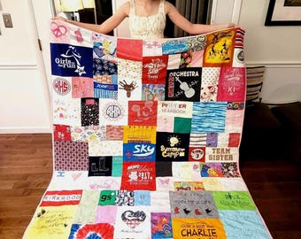 Puede incluir: Una colcha de retazos colorida hecha de camisetas. La colcha está hecha de cuadrados de diferentes colores y patrones, con algunos cuadrados que presentan texto y logotipos. La colcha está siendo sostenida por una persona con un vestido blanco.