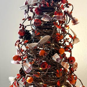 May include: A decorative, conical tree made of woven brown branches, adorned with orange and red berries, small orange pumpkins, pinecones, and plaid ribbons. The tree is lit with small, warm white lights, creating a festive autumn display.