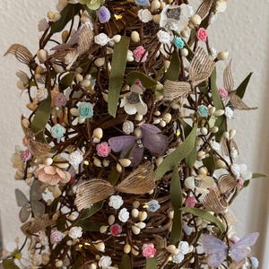 May include: A decorative, cone-shaped tree made of woven brown branches, adorned with small white berries, pastel-colored flowers, green leaves, and butterfly accents. The tree is lit with small, warm white lights.