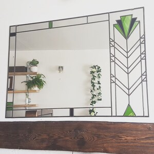 Op de afbeelding: Een grote, rechthoekige spiegel met een zwart metalen frame en groene glazen accenten. De spiegel is omlijst met een geometrisch ontwerp en heeft een decoratief patroon in de rechterbovenhoek. De spiegel reflecteert een kamer met planten en planken.