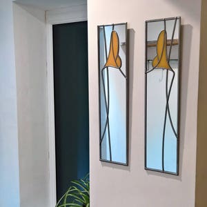 Op de afbeelding: Twee rechthoekige, ingelijste spiegels met een decoratief ontwerp met een geel element van gekleurd glas. De spiegels hangen aan een witte muur.