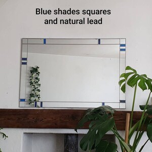 May include: Rectangular mirror with blue square accents and natural lead detailing. The mirror is mounted on a white wall above a wooden mantelpiece. The text "Blue shades squares and natural lead" is above the mirror.