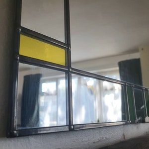 May include: A rectangular mirror with a geometric design. The mirror features a black frame with sections of clear glass, yellow, and green glass. The design is reminiscent of stained glass.