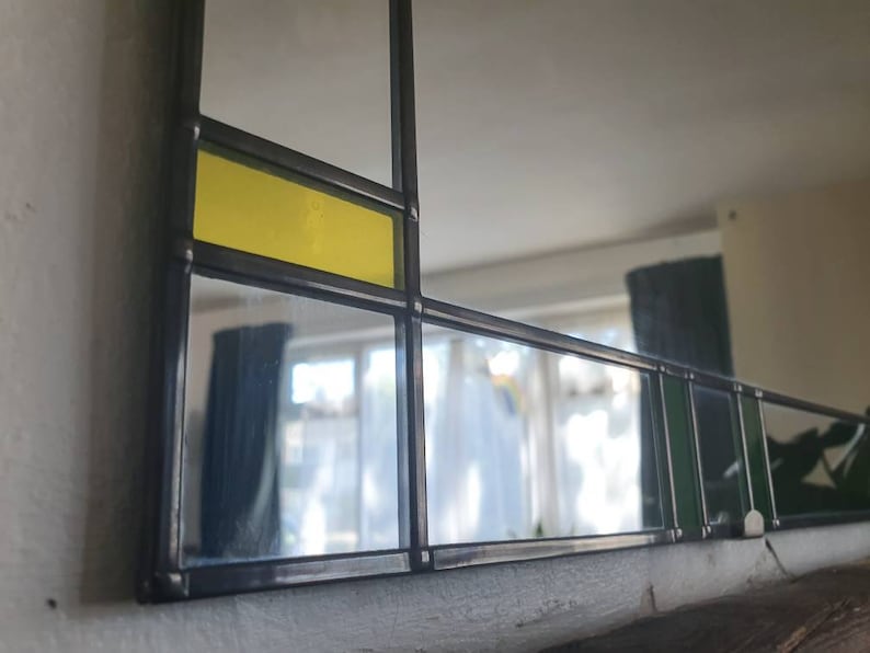 May include: A rectangular mirror with a geometric design. The mirror features a black frame with sections of clear glass, yellow, and green glass. The design is reminiscent of stained glass.