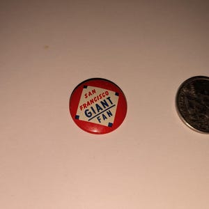 May include: A red San Francisco Giants fan button with white text and a blue border. A US quarter coin is next to the button. The button is a sports collectible.