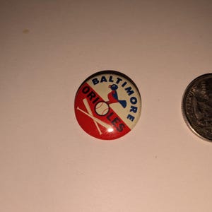 May include: A round Baltimore Orioles button with a red and white design. The button features the team name, a bird logo, a baseball, and crossed bats. A quarter is next to the button.