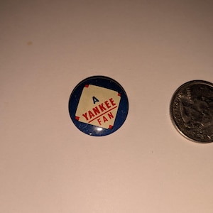May include: A round button with a blue border and the text "A YANKEE FAN" in red and white. Next to the button is a quarter coin. The button is a collectible item.