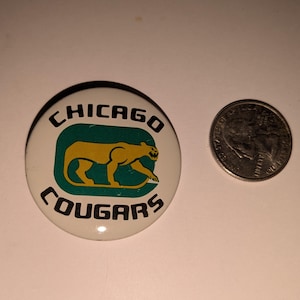May include: A round button with the text "CHICAGO COUGARS" in black lettering. The button features a gold cougar silhouette inside a green rectangle. A U.S. quarter is next to the button for size comparison.