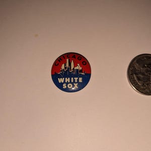 May include: A round Chicago White Sox pin with a red and blue design. The pin features the Chicago skyline and the words "CHICAGO" and "WHITE SOX". A quarter is next to the pin.