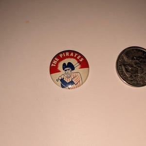 May include: A round button with a red and white background featuring a baseball player holding a bat, with the text "THE PIRATES" in white. A quarter coin is next to the button.