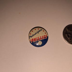 May include: A round Philadelphia Phillies button with the team name in red and white, a baseball, and the city name. The button is next to a quarter.