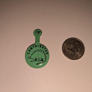 May include: A green pin with the word "CONTRIBUTOR" encircling a black dinosaur illustration. The pin has a small stem at the top. A US quarter is next to the pin for size comparison.