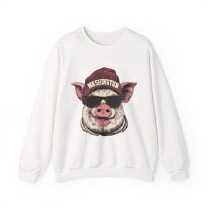 May include: A white long-sleeve sweatshirt featuring a graphic print of a pig wearing black sunglasses and a maroon beanie with the word WASHINGTON printed in white capital letters across the front of the hat.