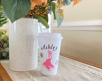 May include: White to-go cup with a pink ice skate design and the name "Ashley" written in black cursive. The cup is sitting on a beige table runner.