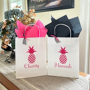 May include: Two white gift bags with pink tissue paper and black tissue paper. The bags have pink pineapple designs and the names "Charity" and "Hannah" printed on them.