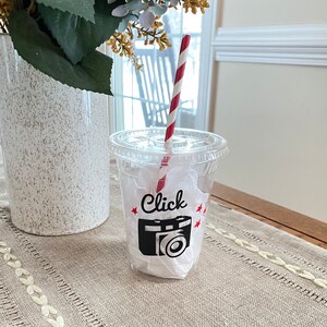 May include: A clear plastic cup with a lid, a red and white striped straw, and a black camera graphic with the word "Click" and red stars. The cup is filled with white paper and sits on a beige tablecloth.
