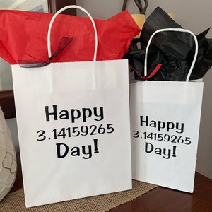 May include: Two white paper gift bags with handles. The bags are decorated with black text that says "Happy 3.14159265 Day!" One bag has red tissue paper inside and the other has black tissue paper inside.