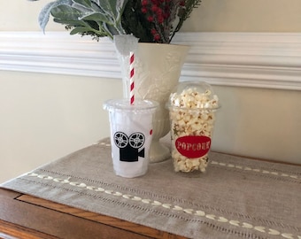 May include: Two clear plastic cups with red and white striped straws. One cup has a black and white film camera design and the other cup has the word "POPCORN" in red letters. The cups are on a brown wooden table with a beige linen runner.