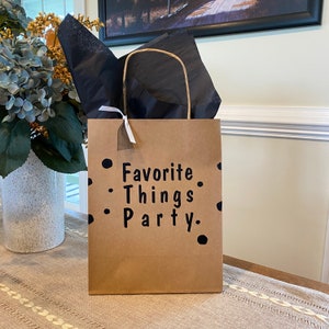 May include: A brown paper gift bag with black polka dots and the text "Favorite Things Party." The bag has a white ribbon tied around the handles.