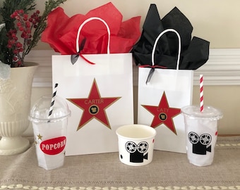 May include: Two white gift bags with red and black tissue paper inside. The bags have a red star with a gold outline and the names "Carter" and "Caty" printed on them. There are two clear plastic cups with black and white striped straws and a white paper cup with a black film camera design. The cups have "Popcorn" and a star design printed on them.