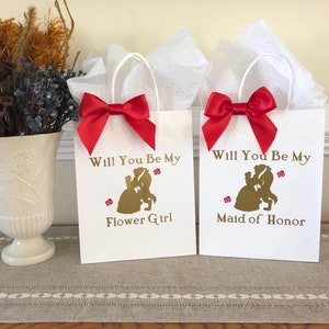 May include: Two white gift bags with red satin bows. The bags have gold lettering that says "Will You Be My Flower Girl" and "Will You Be My Maid of Honor". The bags feature a silhouette of Belle and the Beast from the Disney movie Beauty and the Beast.