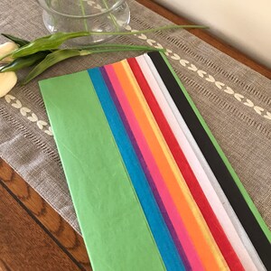 May include: Assortment of tissue paper in various colors, including green, blue, purple, pink, orange, yellow, red, white, and black. The tissue paper is arranged in a stack on a wooden surface.