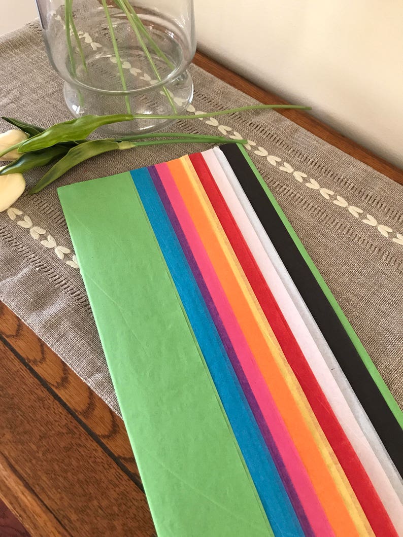 May include: Assortment of tissue paper in various colors, including green, blue, purple, pink, orange, yellow, red, white, and black. The tissue paper is arranged in a stack on a wooden surface.