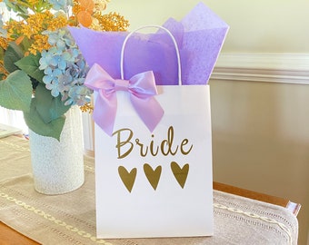 May include: White gift bag with a purple bow and tissue paper. The bag has the word "Bride" printed in gold with three gold hearts below it.